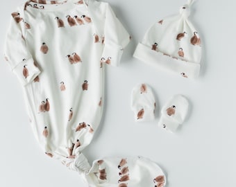 Könnte beinhalten: Ein weißes Baby-Kleid mit einem geknoteten Saum und langen Ärmeln zeigt einen braunen Pinguin-Druck. Eine passende weiße Mütze mit einem geknoteten Oberteil und einem braunen Pinguin-Druck ist enthalten. Zwei weiße Fäustlinge mit braunen Pinguin-Drucken sind ebenfalls enthalten.
