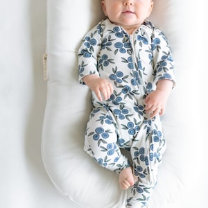 May include: A baby wearing a white and blue long-sleeved sleeper with a blueberry print. The sleeper has a zipper down the front. The baby is lying on a white pillow.