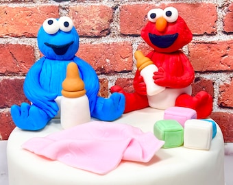 May include: A white cake decorated with a blue Cookie Monster and a red Elmo holding baby bottles. The cake is also decorated with a pink blanket and colorful blocks.
