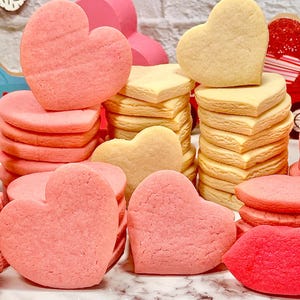 Può includere: Un mucchio di biscotti di zucchero a forma di cuore rosa e gialli. I biscotti sono impilati su una superficie di marmo bianco.