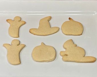Op de afbeelding: Zes onversierde Halloween-koekjes op een witte rechthoekige schaal. De koekjes zijn gevormd als geesten, een heksenhoed, pompoenen en een eekhoorn. De koekjes zijn lichtbruin van kleur en liggen tegen een bakstenen achtergrond.