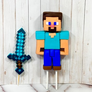 Könnte beinhalten: Ein Minecraft-Themen-Kuchenaufsatz mit einem pixeligen Charakter mit blauem Hemd, braunen Haaren und blauer Hose. Ein blaues und braunes Schwert ist ebenfalls enthalten. Beide sind auf weißen Stäben befestigt.
