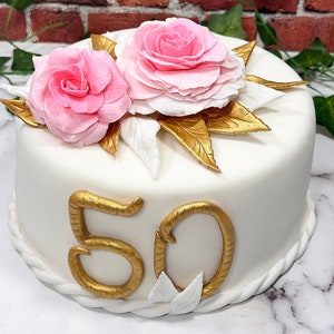 May include: White cake decorated with gold fondant, two pink sugar roses, and gold leaves. The cake is decorated with the number "50" in gold fondant.