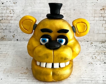 Puede incluir: Una cabeza de oso animatrónico de color amarillo dorado con un sombrero de copa negro, ojos azules y una nariz negra. La boca del oso está abierta, revelando dientes blancos.