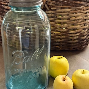Könnte beinhalten: Ein vintage blaues Glasgefäß der Marke Ball Perfect Mason mit einem metallenen Schraubdeckel. Das Glas weist eine geprägte Aufschrift mit dem Text Ball Perfect Mason auf. Das Glas steht neben zwei kleinen gelben Äpfeln.