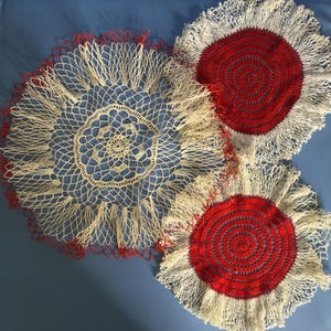 Puede incluir: Tres tapetes hechos a mano en una variedad de diseños. Uno es principalmente de color crema con ribete rojo, mientras que los otros dos presentan un centro rojo con flecos color crema. Los tapetes están dispuestos sobre una superficie azul.