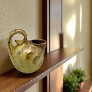 Könnte beinhalten: Eine Keramikkrug mit einem einzigartigen Griff und Ausguss. Der Krug hat einen bauchigen Körper und einen gebogenen Griff. Die Farbe ist eine Mischung aus Gelb, Braun und Beige. Der Krug steht auf einem Holzregal.