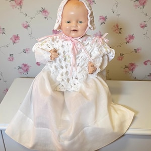 Puede incluir: Una muñeca bebé vintage con la cara pintada, que lleva un chal de ganchillo blanco con cintas rosas y un gorro a juego. La muñeca está vestida con un vestido largo blanco, evocando una estética clásica y nostálgica.