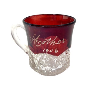 Puede incluir: Una taza de vidrio transparente con una parte superior de vidrio rojo y un patrón de vidrio cortado. La taza tiene un asa y las palabras "Mother 1906" están grabadas en el vidrio rojo.
