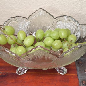 May include: A clear, decorative glass bowl filled with green grapes. The bowl has a scalloped edge and sits on three small feet. The grapes are fresh and ripe, with a natural green color. The bowl is placed on a wooden surface.