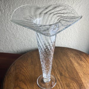 Może przedstawiać: Przezroczysty szklany wazon z szerokim, rozszerzającym się otworem i skręconym, fakturowanym korpusem. Wazon ma okrągłą podstawę i ma około 30 cm wysokości.