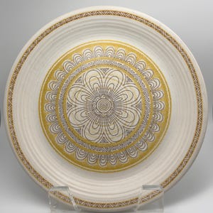 May include: A round ceramic plate featuring a cream-colored base with a central circular motif. The design includes dark brown line-drawn floral patterns surrounded by textured mustard yellow bands and a geometric border along the outer rim.