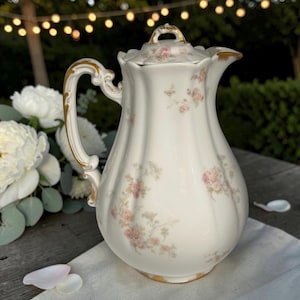 May include: A white porcelain coffee pot with a fluted body and a matching lid. The surface features a delicate pattern of small pink floral sprays and green foliage. The handle, spout, and lid finial are accented with ornate gold trim details.