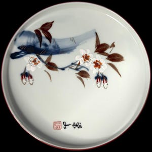 Peut inclure: Une assiette en céramique blanche avec un bord rouge, ornée d'un motif peint à la main. Le motif comprend un oiseau bleu, des feuilles marron et des fleurs blanches avec des centres orange. Des caractères japonais et un sceau rouge sont également présents.