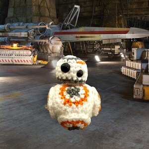 May include: A white, orange, and gray crocheted toy robot with large black eyes. The robot is sitting on a gray floor in front of a large spaceship.