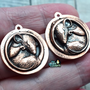 May include: Two copper-coloured metal charms featuring a detailed, three-dimensional design of a sleeping mouse. The charms have a loop at the top for attaching to a necklace or bracelet.