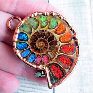 May include: A handcrafted ammonite fossil pendant with a copper-colored frame. The fossil's chambers are filled with vibrant, iridescent gemstones in shades of blue, green, orange, and red. The Glass Cavern logo is visible.