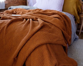 May include: A rust-colored waffle weave throw blanket is draped over a bed with a light purple pillow and a yellow pillow. The blanket is made of a soft, textured fabric and has a relaxed, casual look.