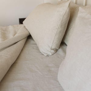 May include: A bed with beige linen bedding, including a duvet and pillows. The pillows are stacked against a white headboard. The bedding has a soft, textured appearance, suggesting a comfortable and inviting bedroom setting.
