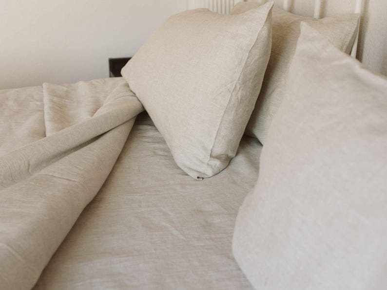 May include: A bed with beige linen bedding, including a duvet and pillows. The pillows are stacked against a white headboard. The bedding has a soft, textured appearance, suggesting a comfortable and inviting bedroom setting.