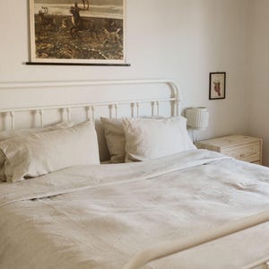 May include: A bedroom scene featuring a white metal bed frame with a linen duvet cover and pillows. A vintage print of deer hangs above the bed. The room has a minimalist aesthetic with a small nightstand and wall art.
