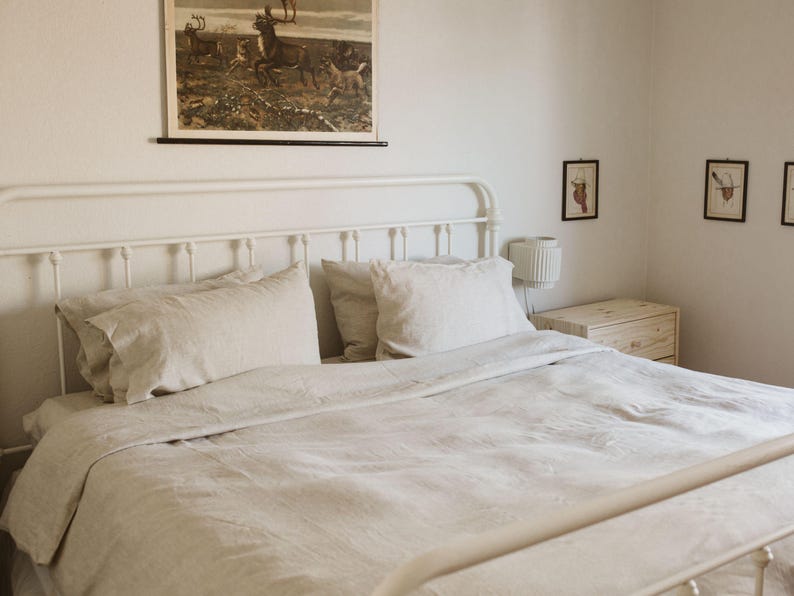 May include: A bedroom scene featuring a white metal bed frame with a linen duvet cover and pillows. A vintage print of deer hangs above the bed. The room has a minimalist aesthetic with a small nightstand and wall art.