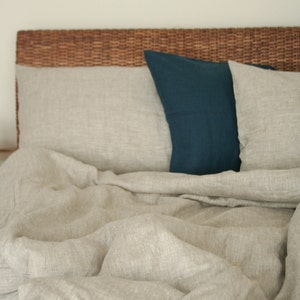 May include: A bed with a woven headboard and neutral-colored linen bedding. The bed has two pillows, one in a deep teal color and the other in a matching neutral tone. The bedding is rumpled, suggesting comfort and relaxation.