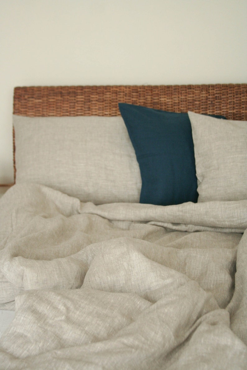 May include: A bed with a woven headboard and neutral-colored linen bedding. The bed has two pillows, one in a deep teal color and the other in a matching neutral tone. The bedding is rumpled, suggesting comfort and relaxation.