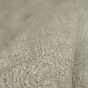 May include: Close-up of a beige linen fabric with a textured weave. The fabric is folded, showing the natural variations in color and texture. The neutral color and soft appearance suggest a natural textile.