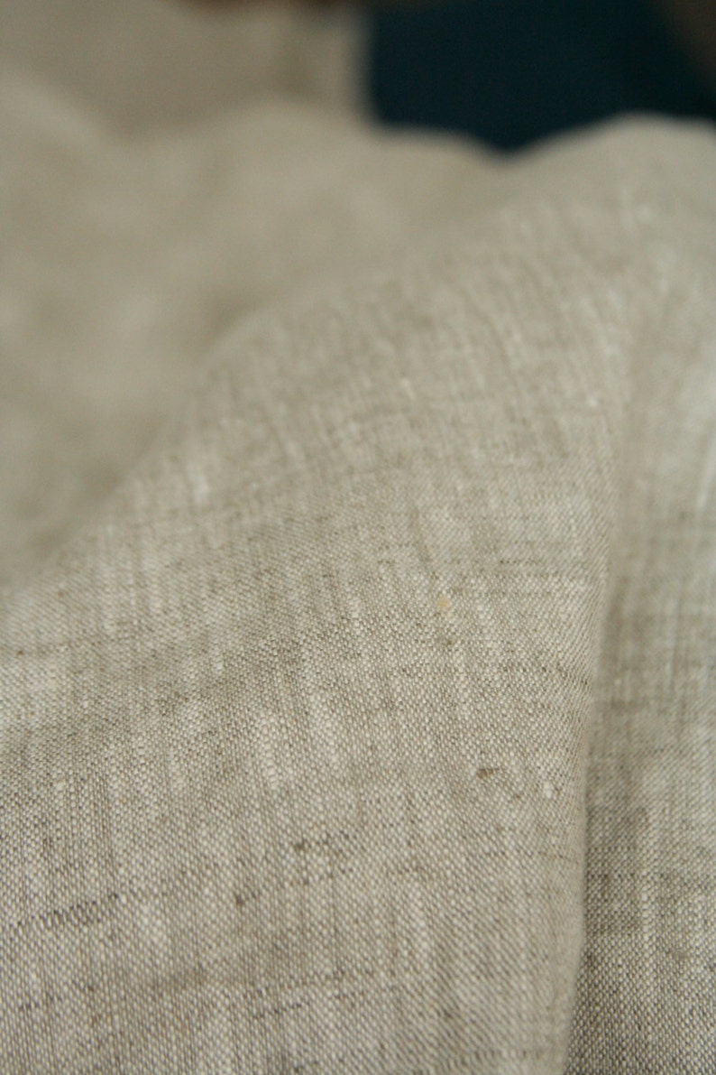 May include: Close-up of a beige linen fabric with a textured weave. The fabric is folded, showing the natural variations in color and texture. The neutral color and soft appearance suggest a natural textile.