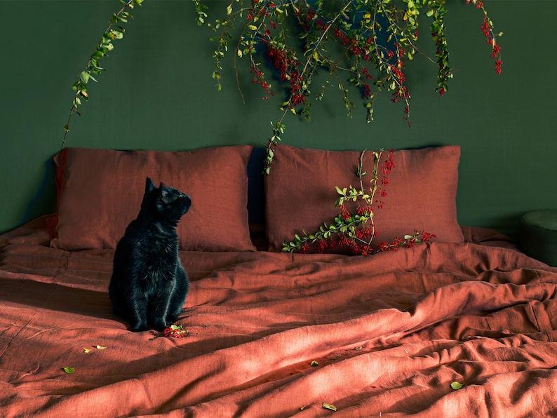 May include: A black cat sits on a bed with rust-colored linen bedding. The bed is against a dark green wall with a leafy branch with red berries hanging above it.