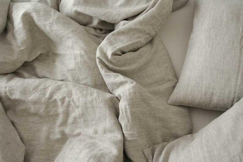 May include: A close-up shot of a bed with a light beige linen duvet cover and pillow. The fabric has a textured appearance with soft folds and wrinkles, suggesting a comfortable and relaxed aesthetic. The neutral color palette creates a sense of calm.