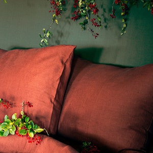 May include: Two rust-colored linen pillowcases on a bed with a green wall in the background. A sprig of green leaves with red berries is on the bed.