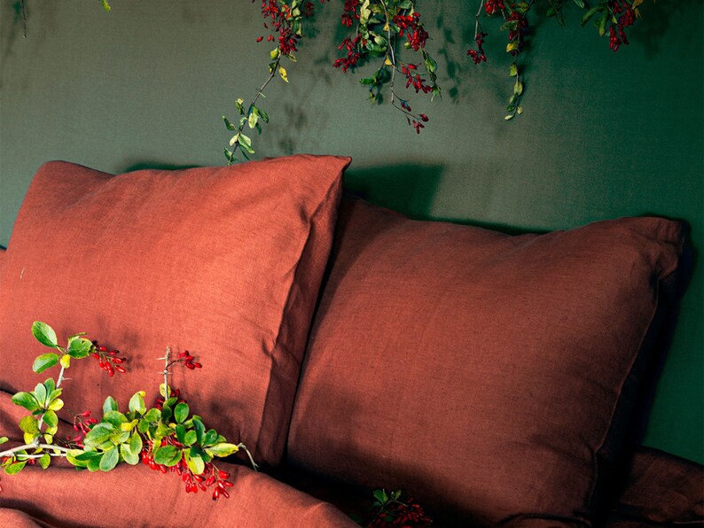 May include: Two rust-colored linen pillowcases on a bed with a green wall in the background. A sprig of green leaves with red berries is on the bed.