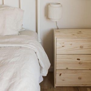 May include: A bedroom scene featuring a bed with white bedding and a three-drawer wooden nightstand. A white lamp with a cylindrical shade is mounted on the wall above the nightstand. The room has a minimalist, neutral aesthetic.