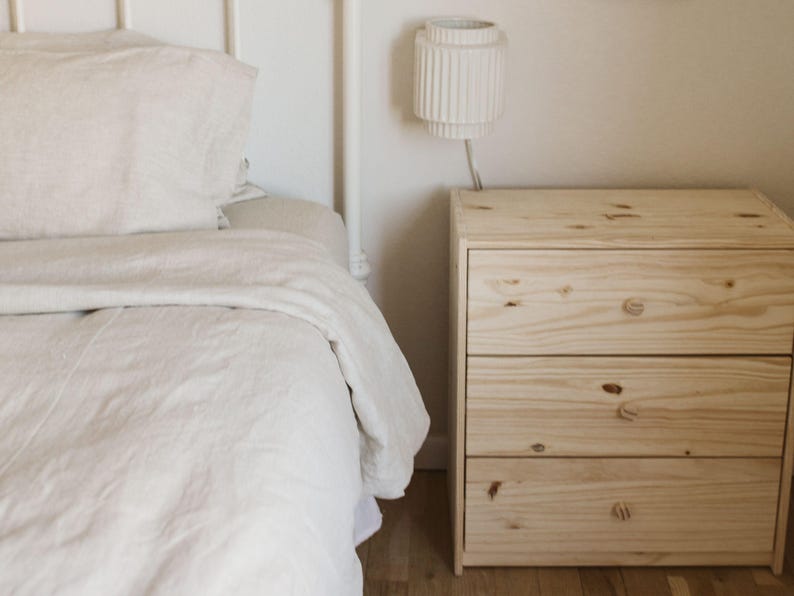 May include: A bedroom scene featuring a bed with white bedding and a three-drawer wooden nightstand. A white lamp with a cylindrical shade is mounted on the wall above the nightstand. The room has a minimalist, neutral aesthetic.