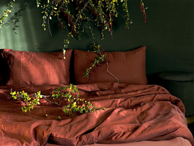 May include: A rust-colored linen bedding set with two pillows and a duvet cover. The bedding is arranged on a bed with a dark green wall behind it. A sprig of greenery with red berries is draped over the bedding.