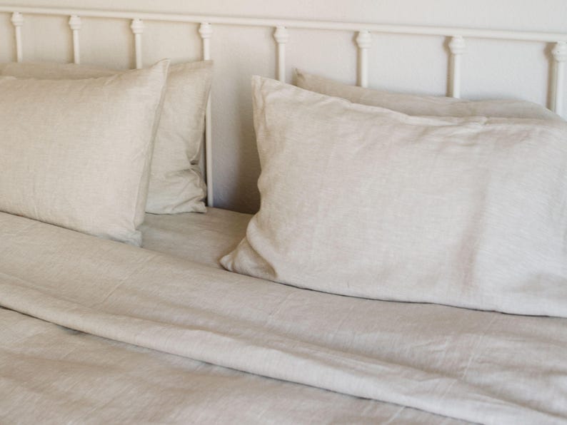 May include: A bed with a white metal headboard and light beige linen bedding. The bedding includes two large pillows and a fitted sheet, creating a soft and inviting aesthetic. The neutral color palette suggests a calming bedroom environment.