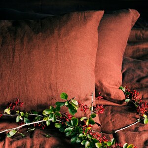 May include: Two rust-colored linen pillowcases on a bed with a sprig of green leaves and red berries.