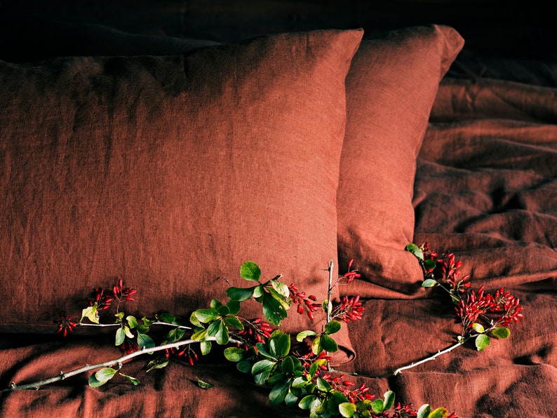 May include: Two rust-colored linen pillowcases on a bed with a sprig of green leaves and red berries.