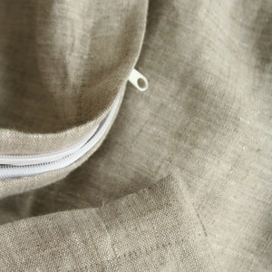 May include: Close-up of a beige linen fabric with a white zipper. The fabric has a textured appearance and is likely part of a pillowcase or bedding. The zipper is partially open, revealing the interior.