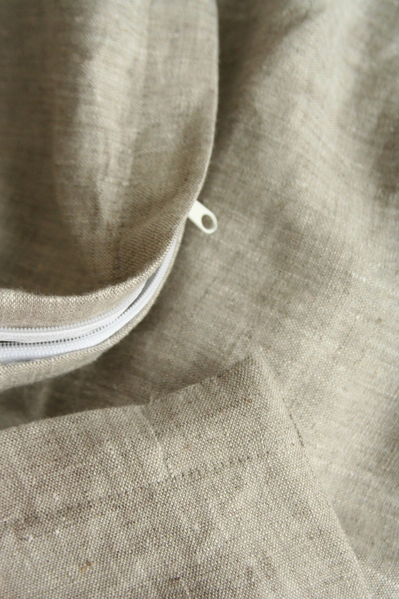 May include: Close-up of a beige linen fabric with a white zipper. The fabric has a textured appearance and is likely part of a pillowcase or bedding. The zipper is partially open, revealing the interior.