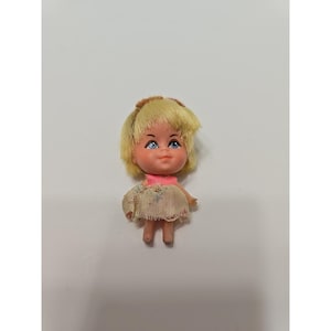 May include: A small doll with blonde hair, blue eyes, and a pink collar. The doll is wearing a light-colored dress. The doll is standing on a white surface. The doll is a vintage toy.