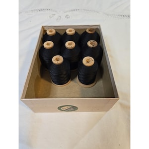 May include: A box filled with nine spools of black thread. The spools are made of wood and have a circular design on top. The box is beige and has a green and white label on the side.