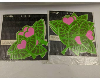 Puede incluir: Dos paquetes de pegatinas vintage con hojas verdes y corazones rosas. Las pegatinas tienen forma de hoja con un corazón en el centro. El embalaje es negro con el texto "Rickie Tickie Sticks".