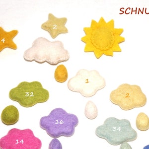 Może przedstawiać: Zestaw filcowych kształtów, w tym chmury, gwiazdy i słońce, w różnych kolorach. Każdy kształt ma wpisaną na nim liczbę. Tekst "SCHNUFFELINIS" jest napisany w prawym górnym rogu obrazu.