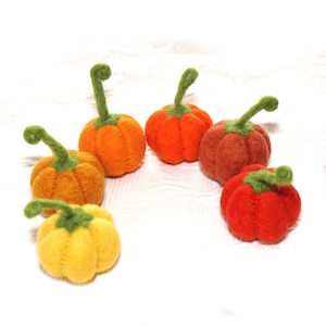 Puede incluir: Una colección de calabazas de fieltro hechas a mano en varios tonos de naranja, rojo y amarillo. Cada calabaza tiene un tallo verde. Las calabazas están dispuestas sobre una superficie blanca, perfectas para la decoración otoñal.