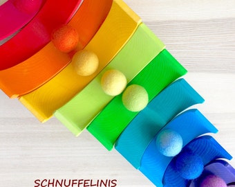 May include: A set of nested wooden rainbow arches in shades of red, orange, yellow, green, blue, and purple, paired with matching felt balls. The text SCHNUFFELINIS is printed in dark red on the left side of the wooden toy set.