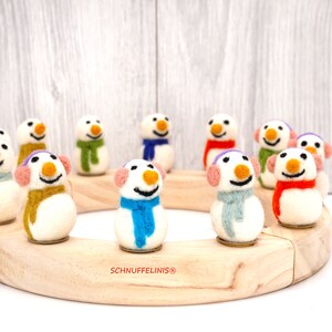 Könnte beinhalten: Ein Adventskalender aus Holz mit Filz-Schneemann-Ornamenten. Die Schneemänner sind in einem Kreis um die Holzbasis angeordnet. Jeder Schneemann hat einen Schal und eine Mütze in einer anderen Farbe. Der Text "SCHNUFFELINIS®" ist auf der Basis aufgedruckt.