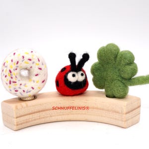 Könnte beinhalten: Eine verspielte Anordnung mit einem Filz-Donut mit bunten Streuseln, einem roten Marienkäfer mit schwarzen Punkten und einem grünen vierblättrigen Kleeblatt. Alle sind auf einem gebogenen Holzsockel mit dem Text "SCHNUFFELINIS®" angebracht.